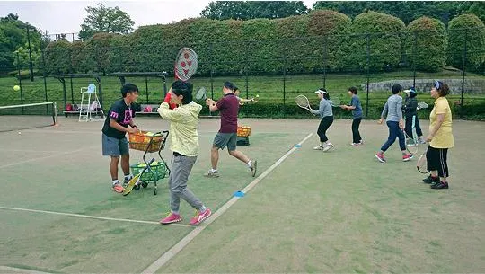 テニススクールで指導する学生と、ラケットを構える人々の様子