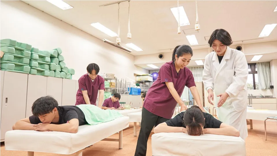 学生たちが講師に教わりながらベッドに横たわる人に施術をしている様子
