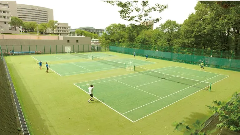 学生たちが周囲が緑に囲まれたテニスコートでテニスをしている様子