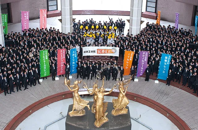 金色の銅像が飾られた吹き抜けのエントランスホールにスーツ姿の大勢の学生が集まり、日本工学院の横断幕や各カレッジの上りを掲げている様子を鳥観撮影した画像