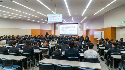 スーツ姿の学生たちが講義室で前方のスクリーンを見ながら説明を受けている様子