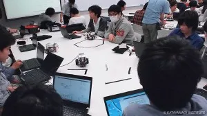 大きなテーブルに10人以上が集まり、プログラムを思案している様子