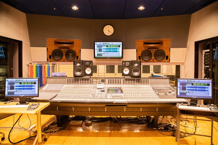 プロ仕様の音響ミキサーが設置された大きなテーブルを中心に音響機器やディスプレイが並び、奥に収録ブースが見える画像