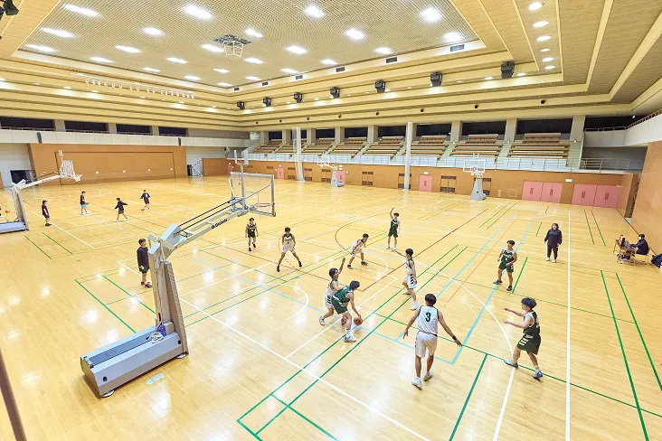 階段状の観客席を持つ館内でバスケットボールの試合をする生徒達