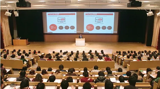学生たちが広い講堂でスクリーンを使った講話を聞く様子