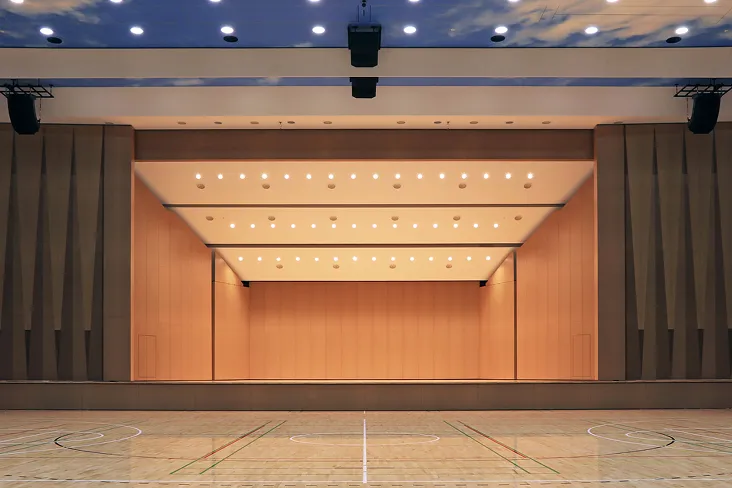 バスケットコートなどのラインが引かれた体育館の床面の奥にモダンなステージがある画像