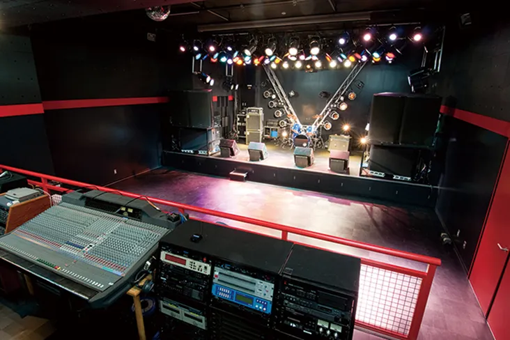 観客席と赤い柵で仕切られたミキサーブースからたくさんの照明や機材が設置されたライブステージを臨む画像