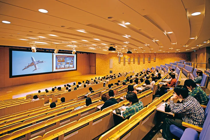 ゆるくカーブする階段型のテーブル付き座席と巨大なスクリーンが2枚備えられたステージで講義が行われている風景