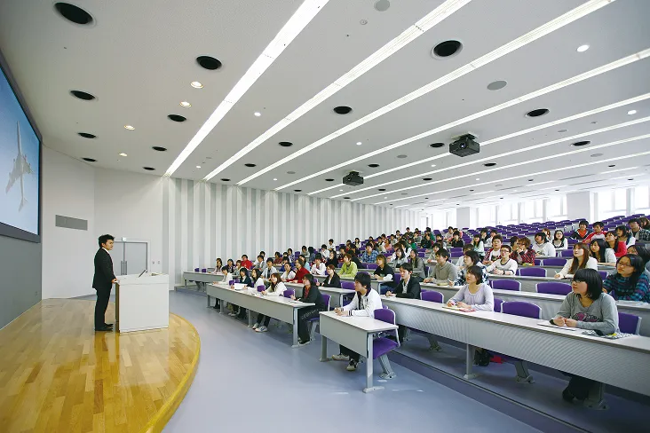 巨大なスクリーンと演台を備えたステージと階段状に講義用デスクの座席が設置されている講義室の授業風景