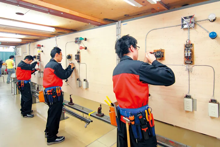 壁に見立てた木の板に設置されたコンセントなどの配電機器に工事を施す実習の様子