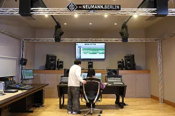 大型ディスプレイを中心にデジタル録音用機器が並んだ室内で指導を受ける生徒の様子