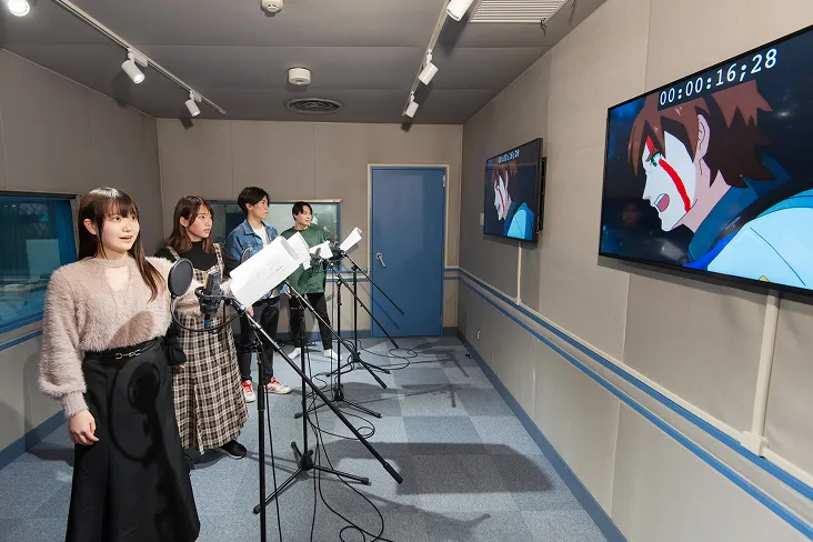 壁に掛けられたディスプレイの映像と台本を見ながらアニメのアフレコをする生徒達