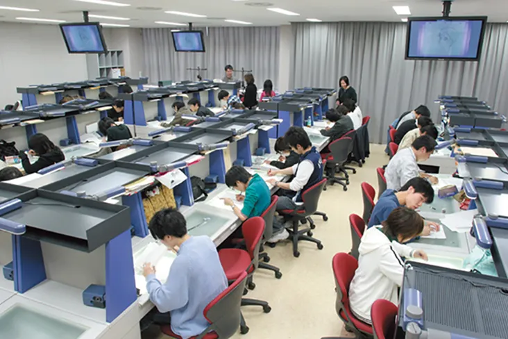 トレース台を兼ねた天板に資料を差し込む物入とデスクライト付きの棚が付いた専用デスクにつき、集中して作業している授業風景