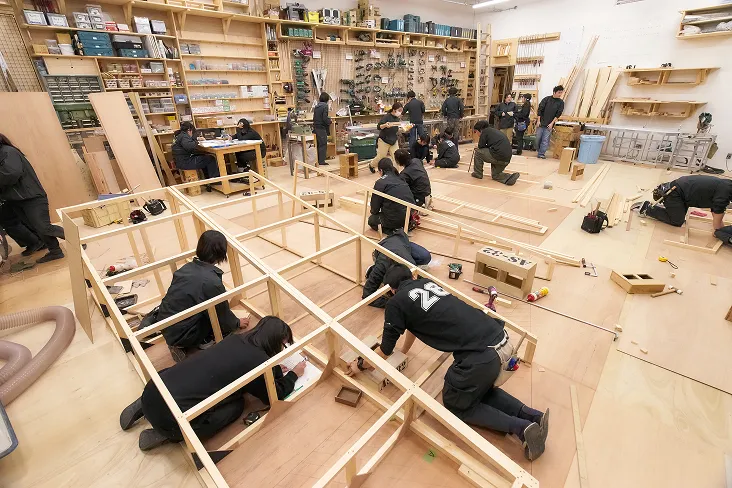 つなぎを着た生徒達が木材と格闘して大型の製作物を作成している風景