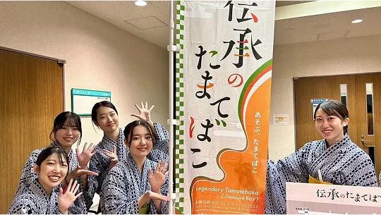 「伝承のたまてばこ」ののぼり旗と一緒に記念撮影をする学生たち