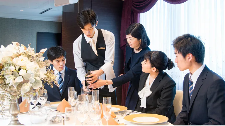 学生たちがテーブルセッティングされた環境で講師からサービスについて学ぶ様子