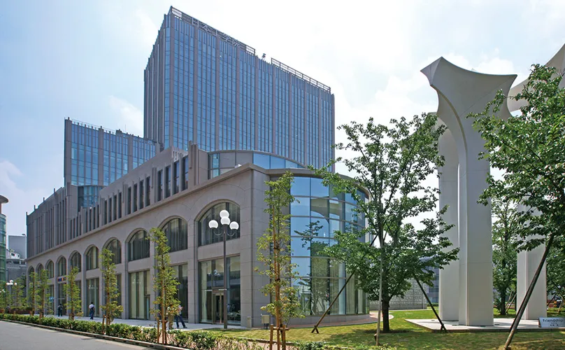 一部に高層階を持つ美術館かホテルのような外観の建物と、街路樹や芝生の豊富な緑の中にオブジェが見える画像