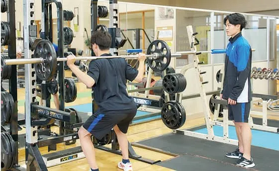 トレーニングマシンを使用している男性と、後ろから見守っている男性の様子