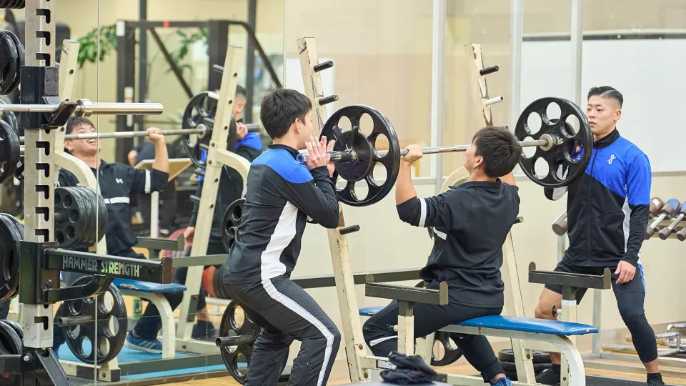 座って行うバーベルを使用したトレーニングをサポートをしている男性2人の様子