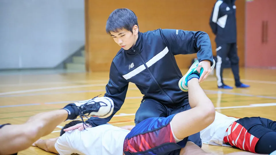 体育館の床に寝そべってのトレーニングをサポートしている様子