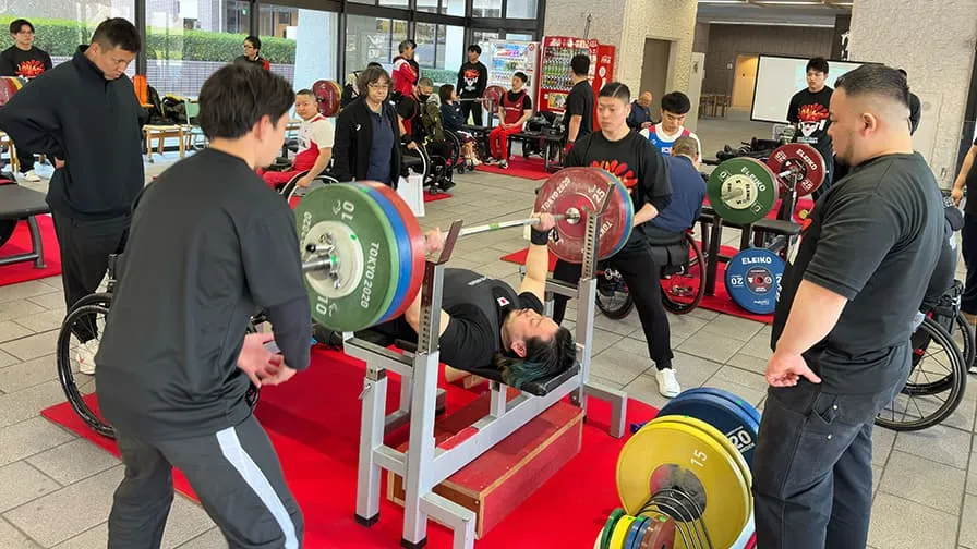 専用の台に仰向けになり、重量プレートがいくつもついたバーベルを持ち上げている選手を囲むようにしてサポートについている様子