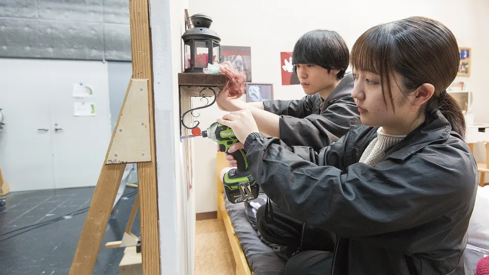美術セット用に電動ドリルで壁に棚をつける学生2人