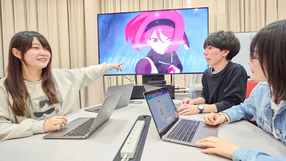 アニメキャラクターが映るモニターに関して、ノートパソコンを囲んで会話する3人の学生