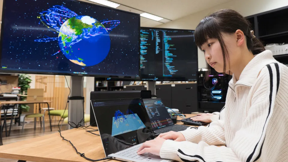学生がノートパソコンで作業し、背後の大型モニターに地球の3DCGが映し出されている