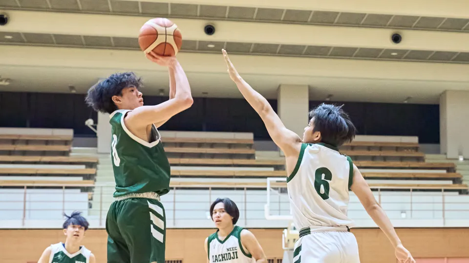 体育館でバスケットボールの試合中、緑のユニフォームの選手がシュートを放ち、白のユニフォームの選手がブロックに跳ぶ。