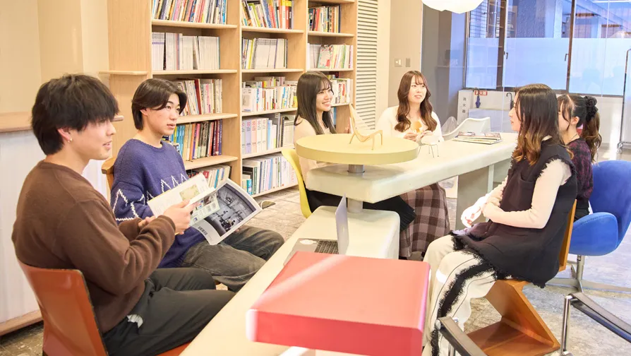 本棚のある教室で雑誌を手に話し合う学生たち