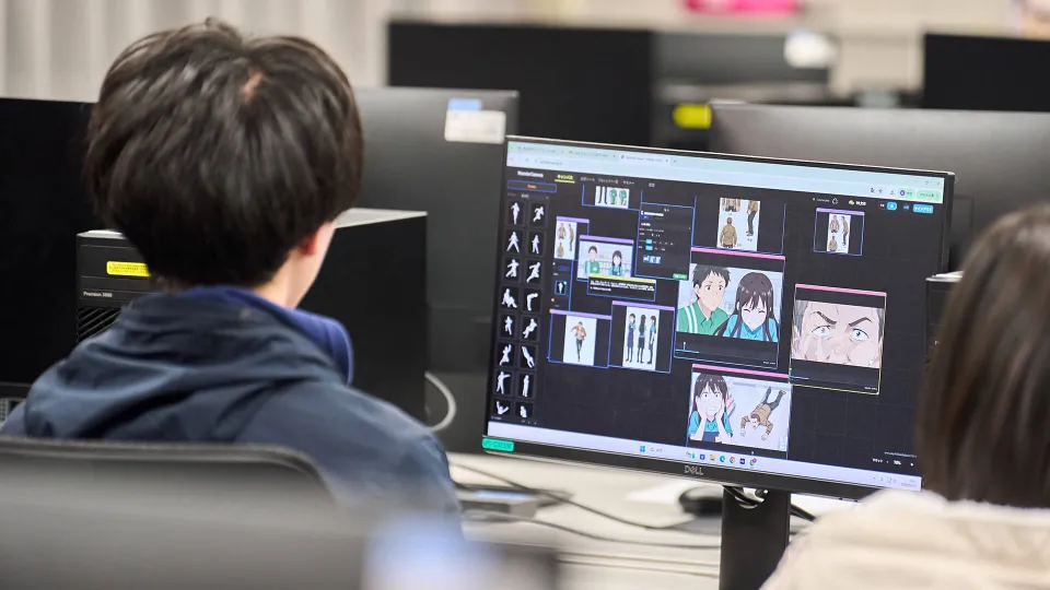 アニメのキャラクターたちが映し出されているパソコンのモニターを見る学生
