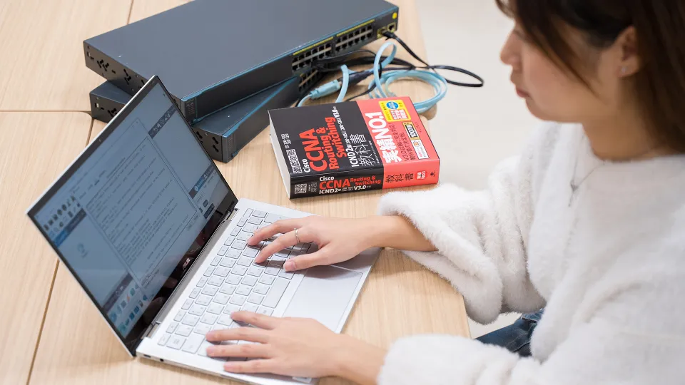 CCNAの参考書とネットワーク機器の横で、ノートパソコンを操作する学生
