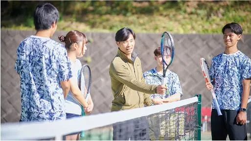 学生たちがラケットを持ち、テニスコートのネットの前で指導を受けている様子