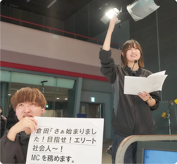 スタジオで台本を手にライトの指示を出す学生と、カンペを持つ学生