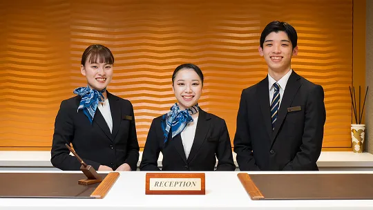 受付カウンターの中に並び、笑顔でお出迎えの姿勢を取るホテルの制服を着た生徒達