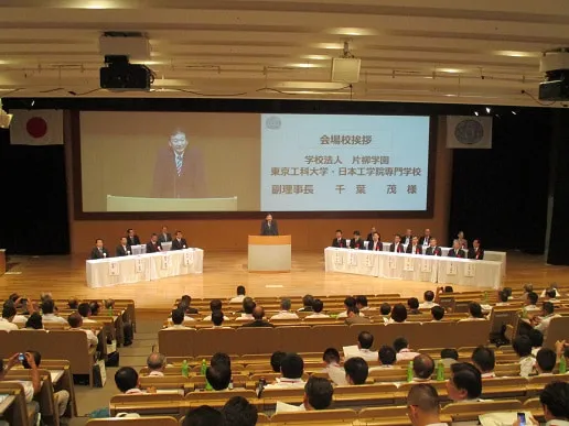 来賓と参加校責任者たちが並ぶ壇上の中央で演台に立ち、挨拶をする理事長