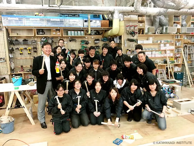 アトリエの一画に工具を持って集まった学生たちと、工具と大きな接着剤を持った理事長で記念撮影