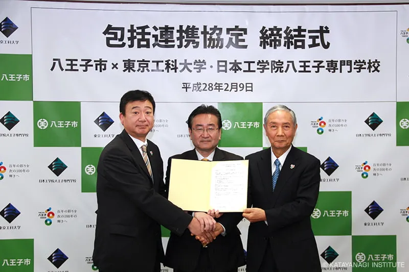 包括連携協定締結式と団体名、日付が掲示された八王子市のバックパネルの前で締結書を持ち記念撮影する3団体のトップ