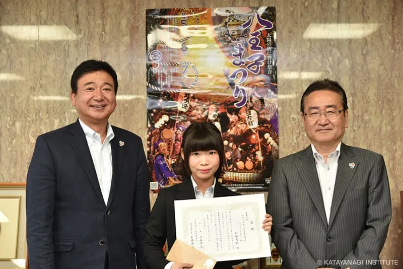 採用されたポスターの前で賞状を持った受賞学生と八王子市市長と共に記念撮影