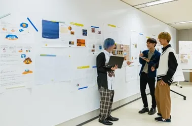 たくさんの資料が貼られた壁の前で立ったまま相談をしている生徒達