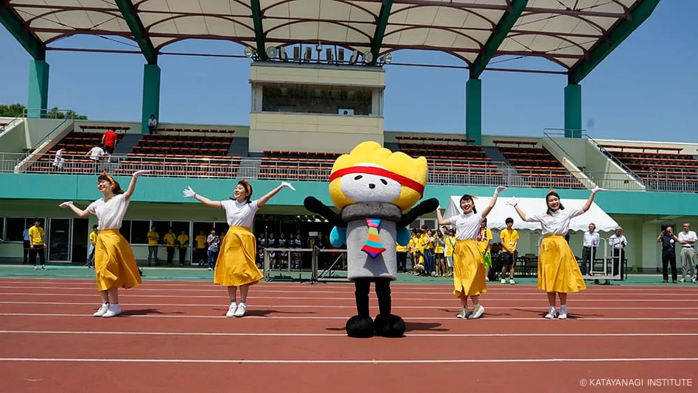 赤い鉢巻きを巻いたかまトゥが黄色のスカートを履いた学生達と両手を広げて踊っている様子