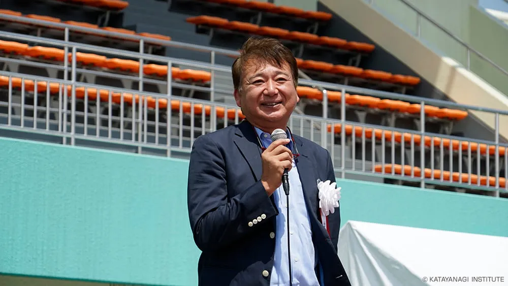 胸章をつけ、風に吹かれながらにこやかに挨拶をする校長