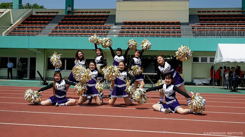紺と白のユニフォームに金銀混色のポンポンを持って決めポーズをするチアチーム