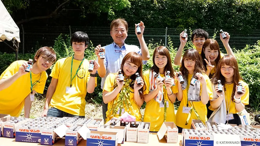 黄色のTシャツの実行委員たちが栄養ドリンクを配るブースにて、ドリンクの瓶を持ってポーズ