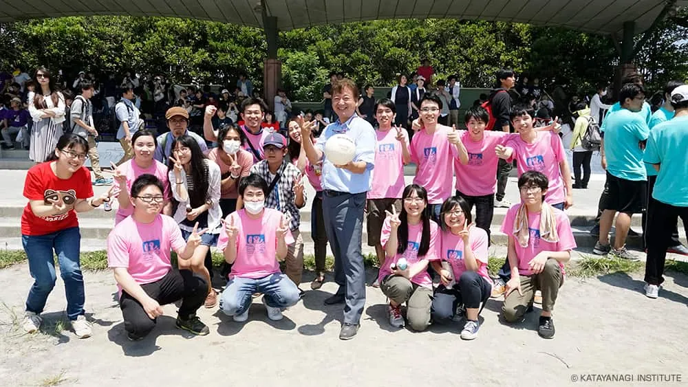 ピンクに青のフロントプリントのTシャツの学生たちがピースをする中心で、ボールを持って片足を前に出したポーズで記念撮影