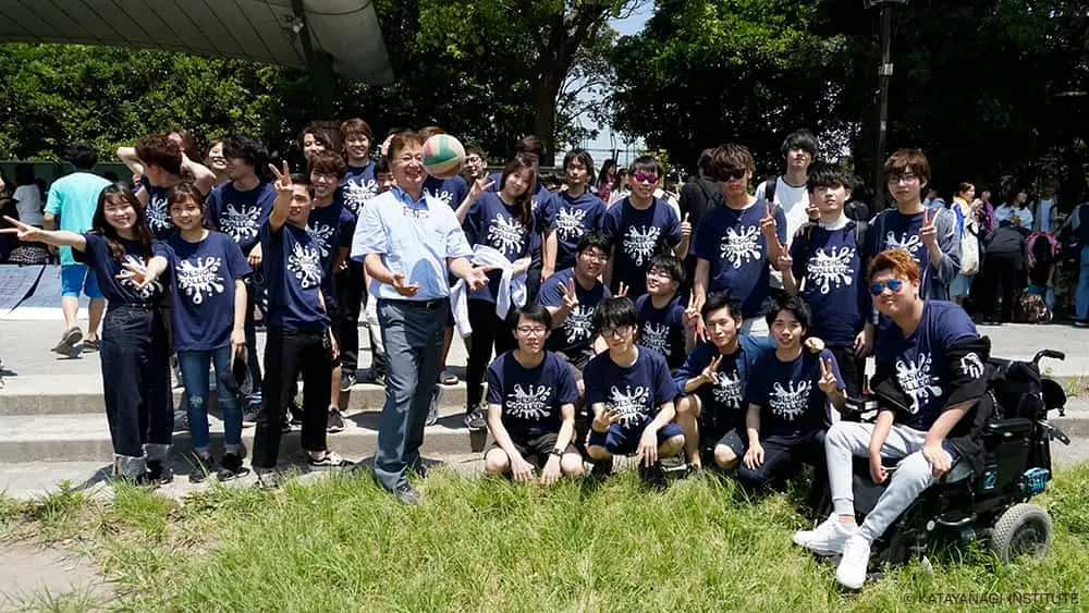 濃紺のTシャツを着た学生たちと、カラフルな配色のバレーボールを上へポンポンさせながら記念撮影