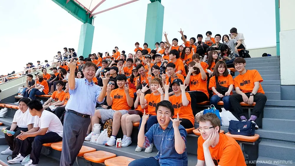 オレンジのTシャツに黒で勉強！！とプリントされた、ユニークなTシャツの留学生たち。階段状の観客席で流行のポーズで1枚