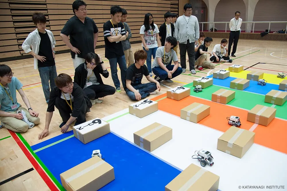 色分けされた6コースに、等間隔に置かれた段ボール箱を認識、回避してゴールへ向かう機体達と、笑顔ではあるが緊張した様子で見守る学生たち