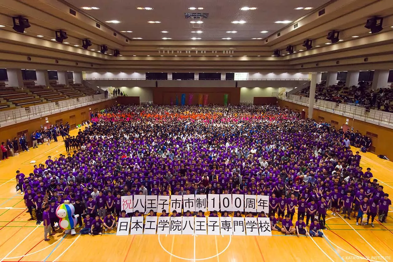 祝 八王子市市制100周年 日本工学院八王子専門学校と1文字ずつ書かれたプラカードを持ち、全学生がアリーナに集合した風景を高い場所から撮影した記念写真