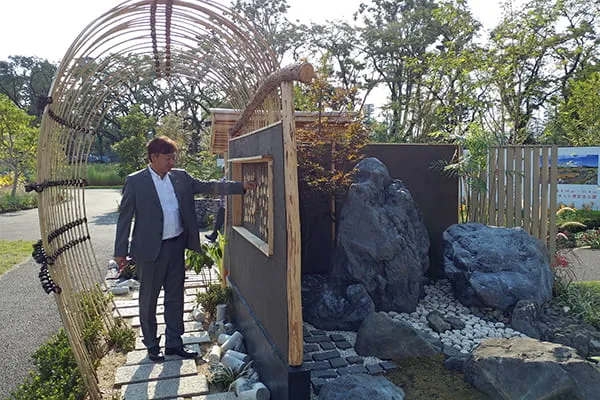 竹材のアーチが架けられた脇道を歩く校長と、壁の向こうに見える大きな庭石の庭園